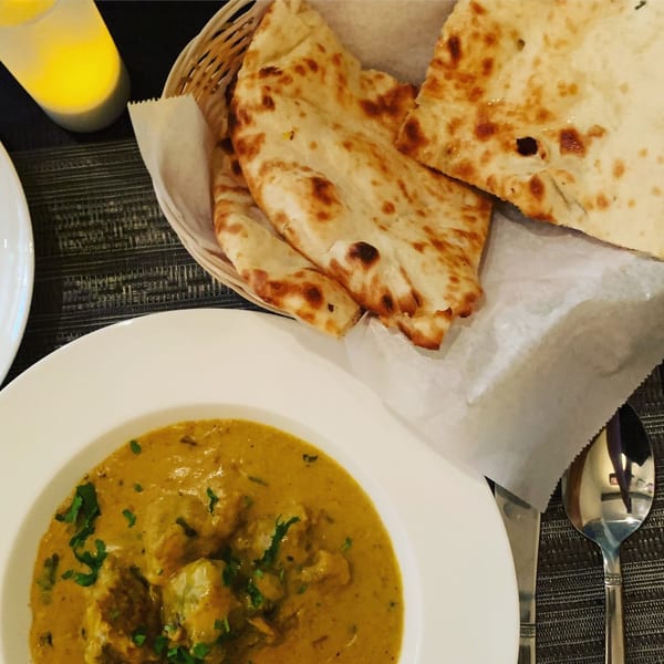 Indian curry with naan bread in a basket, a spoon, and a candle on a dark placemat.