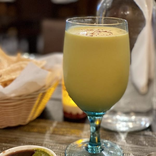 A mango lassi in a wine glass with saffron threads, next to chutneys and a water bottle.