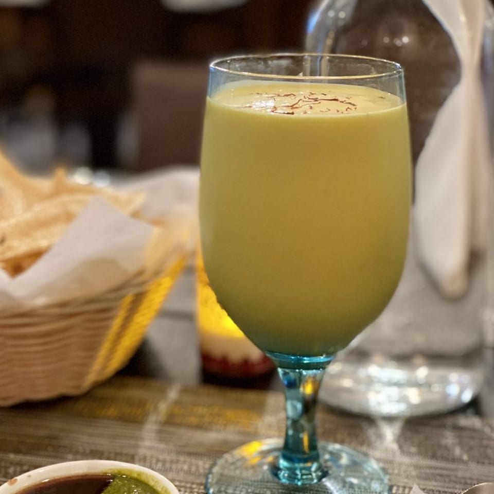 A mango lassi in a wine glass with saffron threads, next to chutneys and a water bottle.