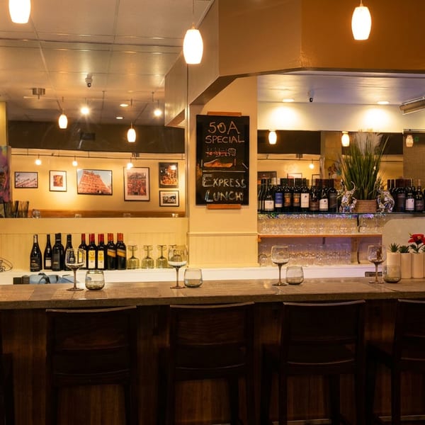 A dimly lit restaurant bar with wine bottles, glasses, and decorative plants under warm pendant lights.