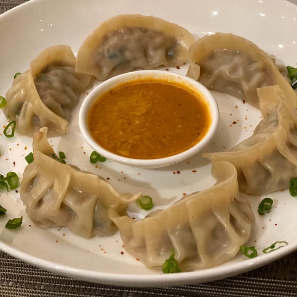 A plate of steamed momos arranged in a circle around a small bowl of orange dipping sauce.