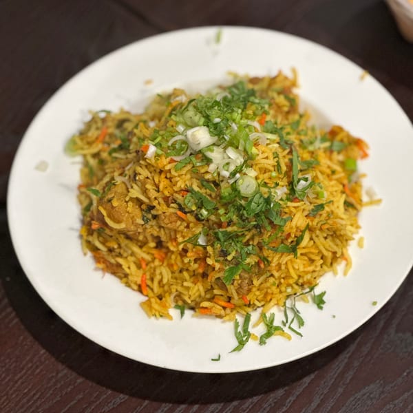A white plate of chicken biryani topped with fresh cilantro and green onions on a dark wooden table.