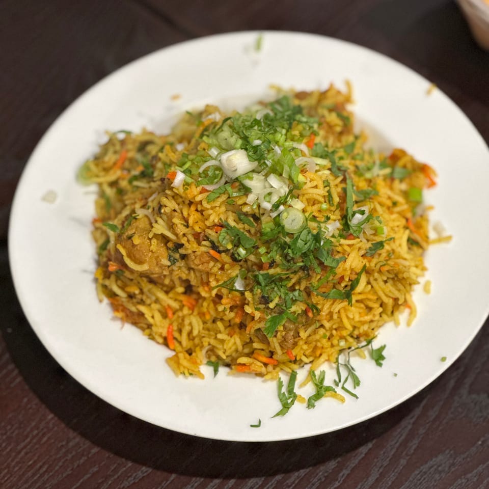A white plate of chicken biryani topped with fresh cilantro and green onions on a dark wooden table.