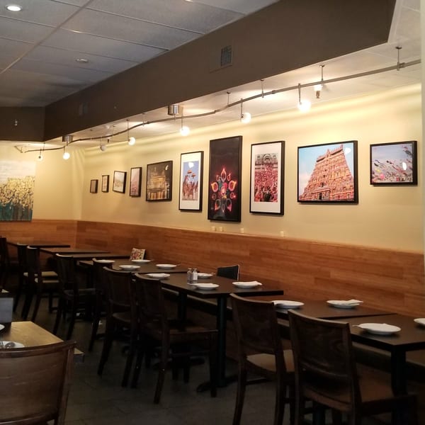Empty restaurant dining room with dark tables, chairs, and cultural artwork on light yellow walls.
